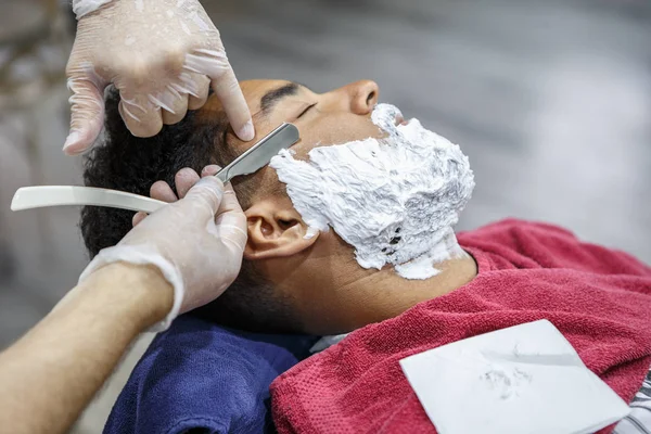 Barber cuts new beard shape on young black man face with sharp razor shaver tool in barbershop salon. Male beauty treatment concept