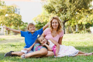 Happy young family having fun in green park at summer day.Attractive blonde mother and her little son and daughter together.Togetherness concept.Unity and love.Pretty white European models outdoors