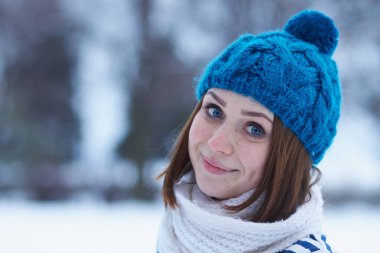 Funny young ginger girl making faces.Blurred background.Beautiful young white girl in warm blue knit winter hat posing outdoor.Winter blurry background out of focus.Place text over blur backdrop 
