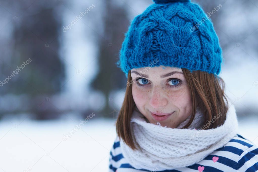 Funny young ginger girl making faces.Blurred background.Beautiful young white girl in warm blue knit winter hat posing outdoor.Winter blurry background out of focus.Place text over blur backdrop 