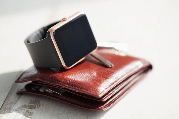 Modern smart watches, brown leather wallet and keys lying on white wooden background. Popular male accessories for everyday usage. Place text or app icon on blank black screen.