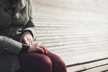 Genç esmer kız açık havada bankta otururken akıllı saatini kullanarak gri paltolu. Bu kişi her zaman sosyal medya ve internet her yerde bağlı.