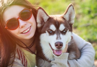 Gülümseyen esmer kız köpeğiyle birlikte açık yeşil parkta oturuyor. Doğanın tadını çıkaran sevimli ve dost canlısı bir çift. Güzel dişi ve pofuduk köpek. 