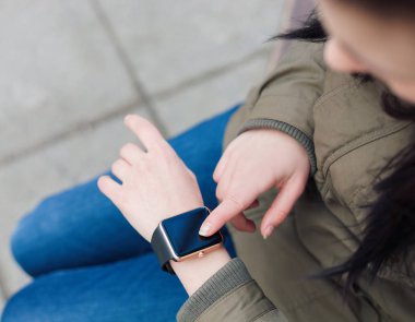 Kız bir bankta oturan akıllı saatini kullanarak parka paltolu. Bu yeni becerikli alet her zaman internet ve sosyal medya ağları ile istediğiniz yerden bağlı kalmak sağlar.