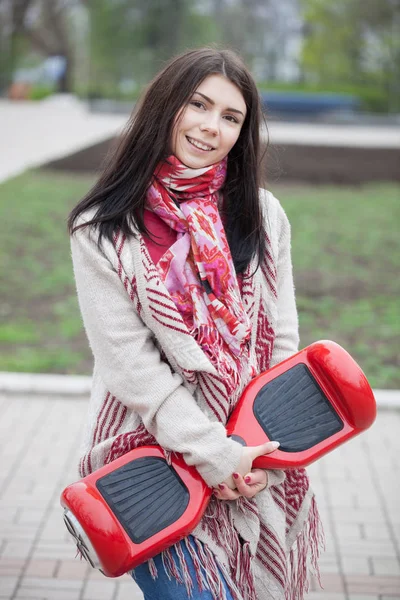 Esmer beyaz girll modern kırmızı elektrikli mini segway ya da vurgulu kurulu scooter parkta yürürken elinde tutan. Popüler yeni becerikli alet. Kız trendy boho tarzı kıyafetler giyiyor.