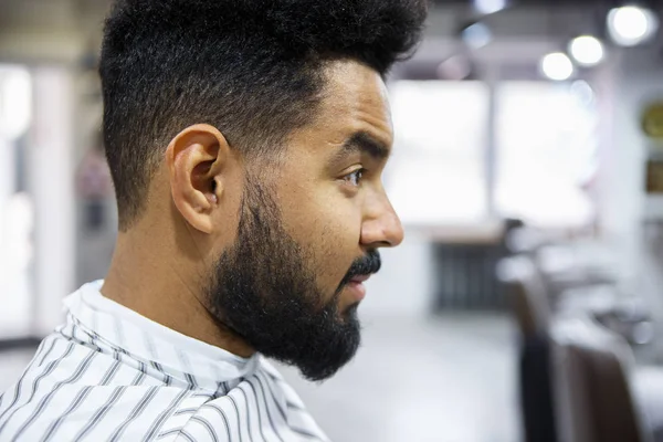 Portrait of handsome young black man with unshaven beard sitting in barbershop waiting for barber to make him pretty haircut and trim beard & mustache hair.Male beauty treatment concept