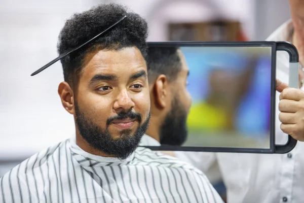 Portrait of black man with comb in curly hair looking in the mirror to the check haircut.Barber showing client his work in barbershop salon.Male beauty treatment concept