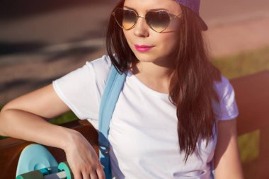 Genç ve güzel esmer kız kalbinde kısa Kruvazör longboard paten güverte ile parkta bankta oturmuş güneş gözlüğü şeklinde