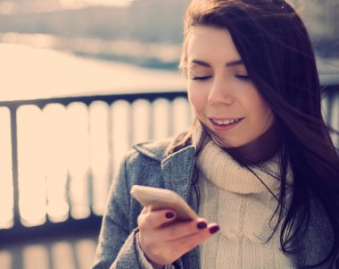 Mutlu bir gülümseme ile dokunmatik ekranlı yeni modern akıllı telefon kullanan bir güzel genç beyaz kadın portresi. Çekici bir modeldir ve güneş parlıyor. 