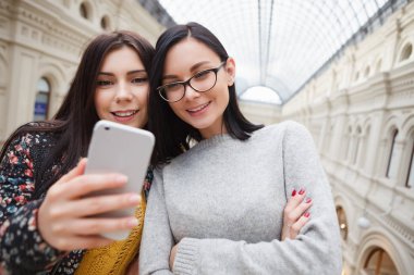 Selfie alışveriş merkezinde alarak iki genç çekici gülümseyen kız. Kadın elinde büyük trendy akıllı telefon. Mobil fotoğraf bir eğilimdir. Modellerde odaklanmak
