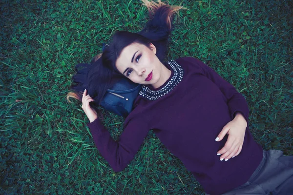 美丽的年轻女孩穿着时尚的玛莎拉毛衣外套躺在绿草地上 面带微笑 模型有红色的口红和明亮的化妆 — 图库照片