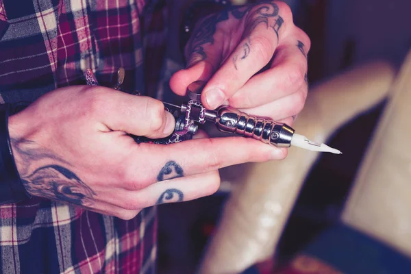 Professional tattoo artist hold tattooing machine in hand.Tattooed hands of tattooist master in beauty studio.Skin art salon essentials,focus on tattoo gun needle.Blue hipster film toning