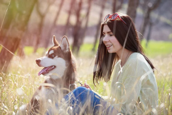 在室外的绿地公园里 一个年轻的黑发女孩和她的哈士奇狗坐在一起 可爱和友善的夫妇享受大自然的美景 漂亮的雌性和毛茸茸的小狗 — 图库照片