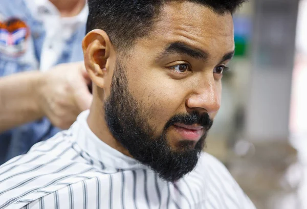 Portrait of unshaven young African man sitting in chair covered with blanket at barbershop salon while barber prepares his hair for cutting.Male beauty treatment process.Cut new haircut & trim beard