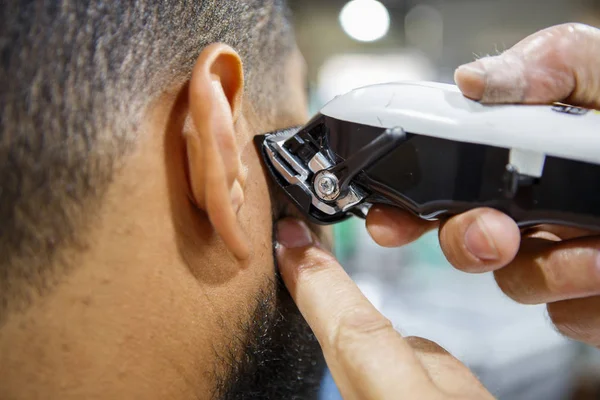 Barber trim African man client with clipper machine in barbershop.Professional trimmer tool cuts beard and hair on young black guy in barber shop salon. Male beauty treatment process in close up