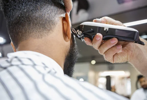 Barber trim hair with clipper on young unshaven black man in barbershop studio.Professional hairdresser cut hair with electric shearer machine on African guy. Male beauty treatment process