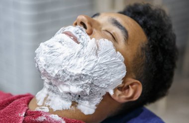 Berberde tıraş için yüzünde beyaz köpük genç siyah adam portresi. İstemci sakal üzerine krema ile. Erkek güzellik tedavi kavramı