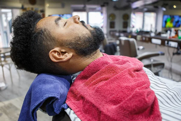 Young black man client lying on barbershop chair waiting for barber to make him new haircut and trim his beard intro shape. Male beauty treatment concept.African guy in hairdresser salon