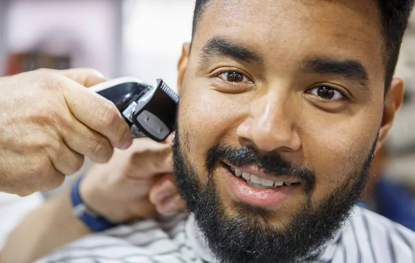 Portrait of happy black man in barbershop.Barber trim clients beard with electric clipper machine in closeup.Male beauty treatment process.Handsome African guy posing at hairdresser salon