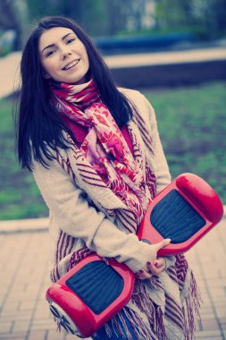 Kadın modeli modern kırmızı elektrikli mini vurgulu kurulu scooter parkta yürürken elinde tutan. Popüler yeni becerikli alet. Kız moda boho tarzı kıyafetler giyiyor.