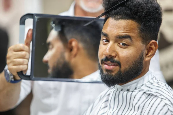 Portrait of handsome black man looking in mirror on his haircut in barbershop studio.Barber hairdresser showing client hair cut in salon.Male beauty treatment concept