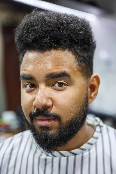 Portrait of young unshaven black man covered with blanket in barbershop salon waiting for new haircut and beard & mustache trimming. Male beauty treatment concept.