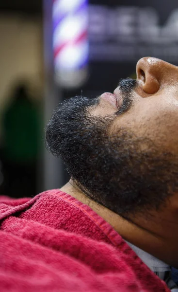 Unshaven black man covered with blanket in barbershop.Client waiting for barber to cut his beard and trim mustach hair.Male beauty treatment concept