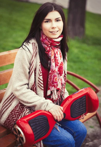 手の現代赤い電気ミニ セグウェイやホバー ボードのスクーターを押しながら緑豊かな公園のベンチに座っている女性モデル 人気のある新しい都市交通 女の子は自由奔放に生きるスタイルのトレンドの服を着ています — ストック写真