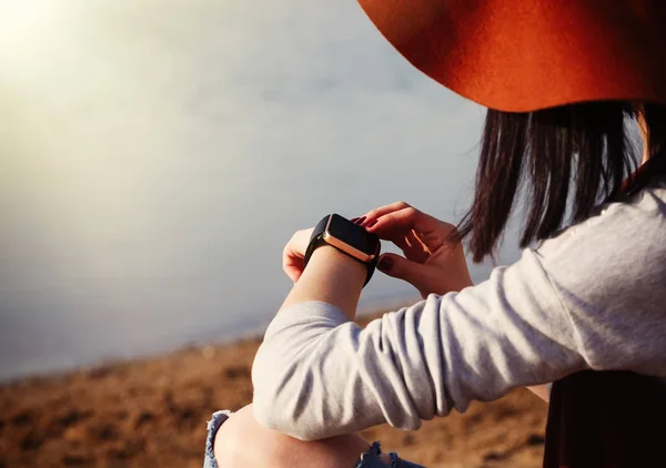 Fashionable brunette woman in boho style clothes using her modern smart watches sitting on the beach. Cute white model, trendy wireless technology that let you always stay connected to internet