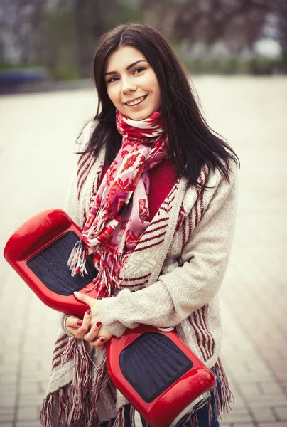 Young brunette white girll holding modern red electric mini hover board scooter in hands while walking in the park. Popular new gadget. Girl is wearing fashionable boho style clothes.