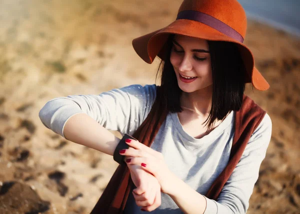 Young fashionable brunette girl in boho style clothes using her modern smart watches sitting on the beach. Cute white model, trendy wireless technology that let you always stay connected to internet