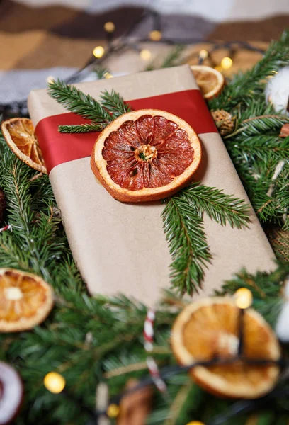 真棒手工制作的圣诞节礼品盒 装饰棕色纸包装 红色丝带 干橘子果包围着松树的花环树枝 背景上的加兰博克灯 — 图库照片