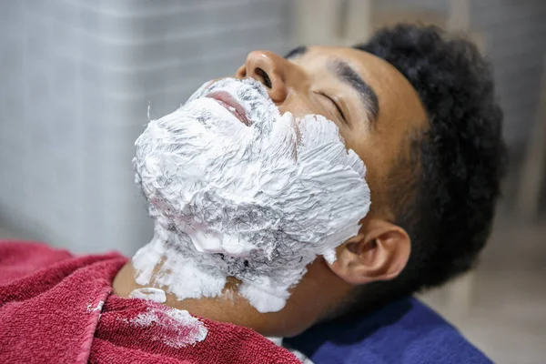 Portrait of young black guy with white foam on face for shaving in barbershop.Client with cream on beard. Male beauty treatment concept