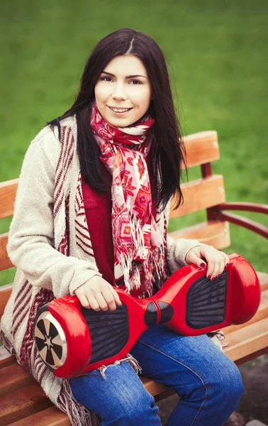 若い白人のブルネットの女の子の手で公園のベンチに座りながらモダンな赤い電気ミニ ホバー ボード スクーターを保持します 人気のある新しい電気都市交通 女の子は自由奔放に生きるスタイルのトレンドの服を着ています — ストック写真