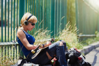 Punk patenci kız trendy kablosuz kulaklıklar açık havada otururken ona Pano montaj yırtık kot pantolon 