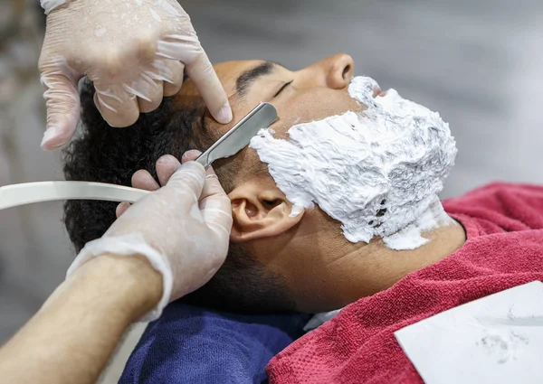 Barber cuts new beard shape on young black man face with sharp razor shaver tool in barbershop salon. Male beauty treatment concept