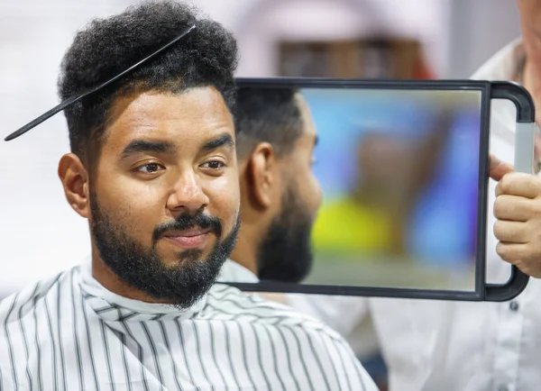 Portrait of black man with comb in curly hair looking in the mirror to the check haircut.Barber showing client his work in barbershop salon.Male beauty treatment concept