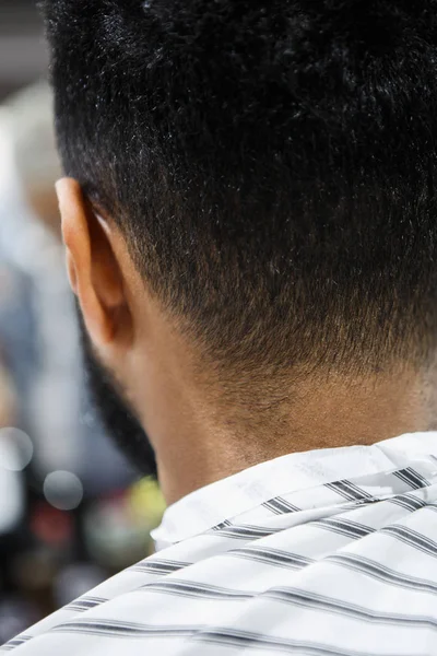 Back of the head and nape of young African man in barbershop. Male beauty treatment process in barber shop.Black guy covered with blanket cuts his hair in salon