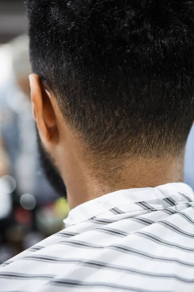 Back of the head and nape of young African man in barbershop. Male beauty treatment process in barber shop.Black guy covered with blanket cuts his hair in salon