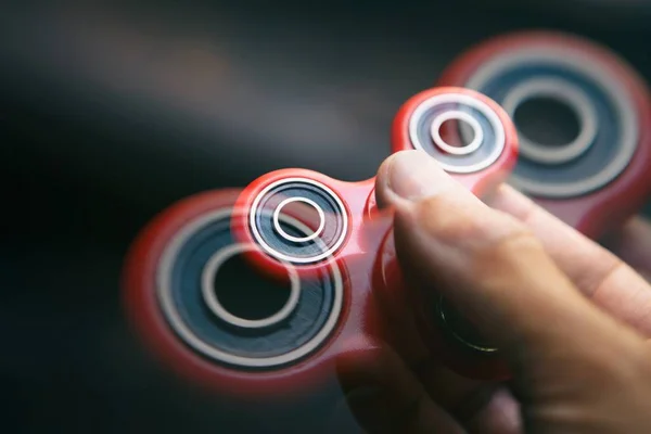 Super popular red spinner device in male hand.Guy holding fidget gadget in hands.Modern spinning toy for youth & adults.Learn cool new tricks & have fun.Red object,dark background