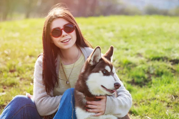 若いブルネットの女の子が屋外の緑の公園で彼女の汚い犬と座って笑っています 可愛くてフレンドリーなカップルが自然を楽しんでいます 可愛い女とふわふわ犬 — ストック写真