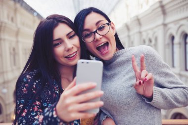 Mutlu genç kız arkadaşlar selfie modern Çift objektifli fotoğraf makinesi akıllı telefon ile alışveriş merkezinde alarak. Komik suratlar, güzel kadın ve trendy yeni smartphone modeli