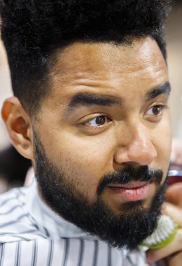 Berber içinde oturan tıraşsız genç siyah adam portresi Kuaför onu yeni şık saç ve sakal ve bıyık saç kesim yapmak bekleyen battaniye ile kaplı. Erkek güzellik tedavi kavramı