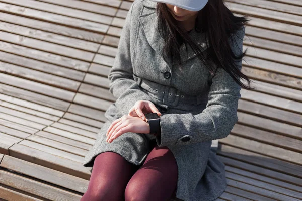 Young brunette girl in grey coat using her smart watch while sitting on the bench outdoors. This person is always connected to social media and internet everywhere.
