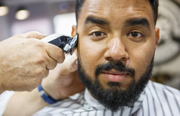 Portrait of young black man being clipped with professional clipper machine in barbershop.African guy makes new haircut and trim beard at barber salon. Male beauty treatment concept