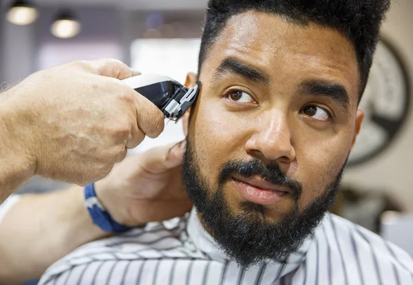 Unshaven young black man being clipped with professional electric shearer machine in barbershop.Male beauty treatment concept.African guy trim beard and mustache at barber shop salon