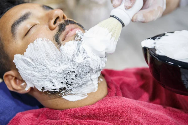 Barber spreads white shaving foam on clients face for accurate shave with razor shaver tool.Young black man get new beard haircut in barbershop salon. Male beauty treatment concept