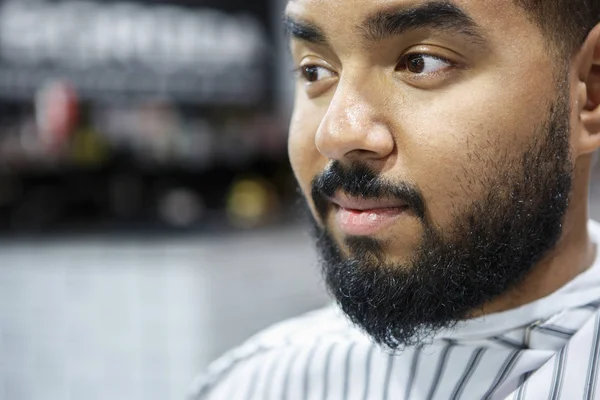 Portrait of unshaven young black man sitting in barbershop chair covered with blanket waiting for barber hairdresser to cut his hair and trim mustache and beard. Male beauty treatment concept