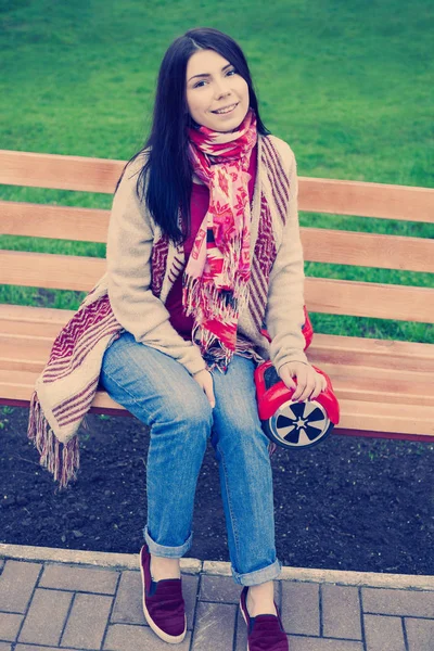 女性モデルは 緑豊かな公園のベンチに座りながら手でモダンな赤い電気ミニ ホバー ボード スクーターを保持します 人気のある新しい都市交通 女の子は自由奔放に生きるスタイルのトレンドの服を着ています — ストック写真