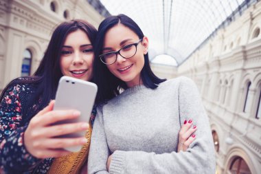 Selfie Çift objektifli fotoğraf makinesi cep telefonu ile alarak çekici esmer kızlar. Modern smartphone becerikli alet, genç ve güzel beyaz modelleri alışveriş merkezinde. Yeni kameralı cihaz